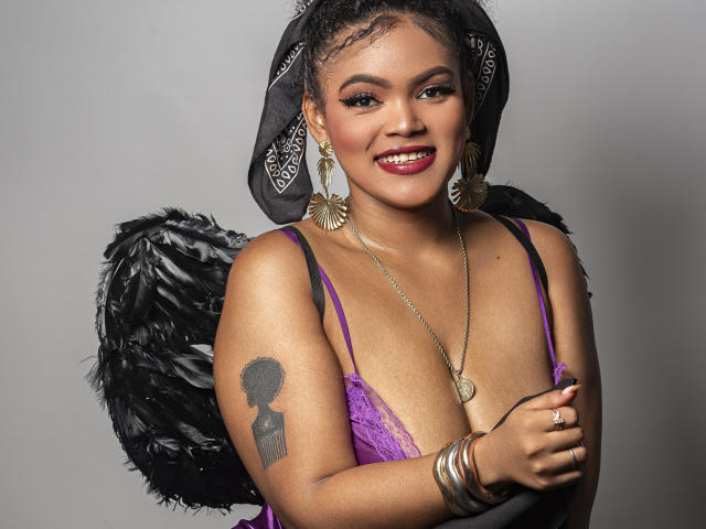 AliceJelanni, con la pelle scura, i capelli ricci, il rossetto rosso, indossa un reggiseno di pizzo viola, ali di piume nere, gioielli dorati e un foulard nero. Tatuaggio visibile sulla parte superiore del braccio, con un'espressione seducente e sorridente..