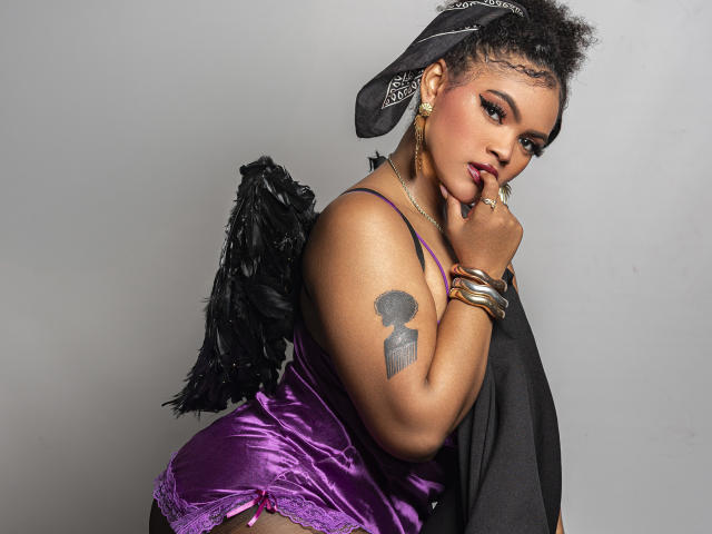 Photograph of AliceJelanni, a curvy Black woman with short, curly hair, wearing a purple satin corset and black wings, touching her chin with her finger. Tattoo on her upper arm. Sexy, confident pose.