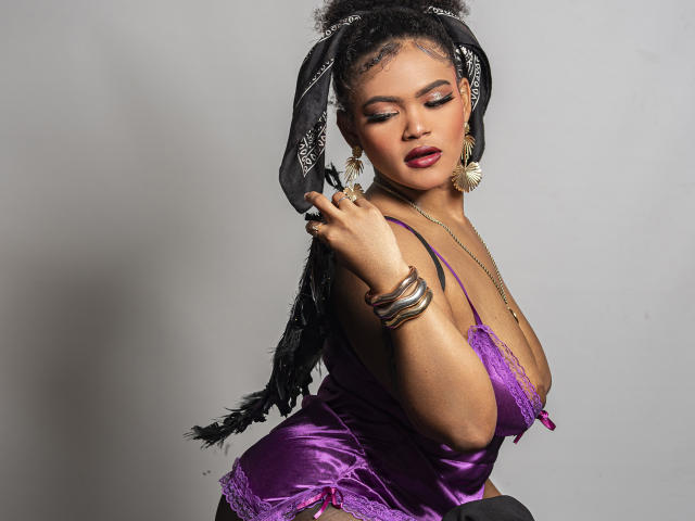 Alice Jelanni, con piel oscura y cabello negro recogido en una coleta, vestía una blusa morada de encaje con escote pronunciado, aretes dorados y brazaletes. Mirando hacia abajo con una expresión seductora, la mano cerca del rostro..