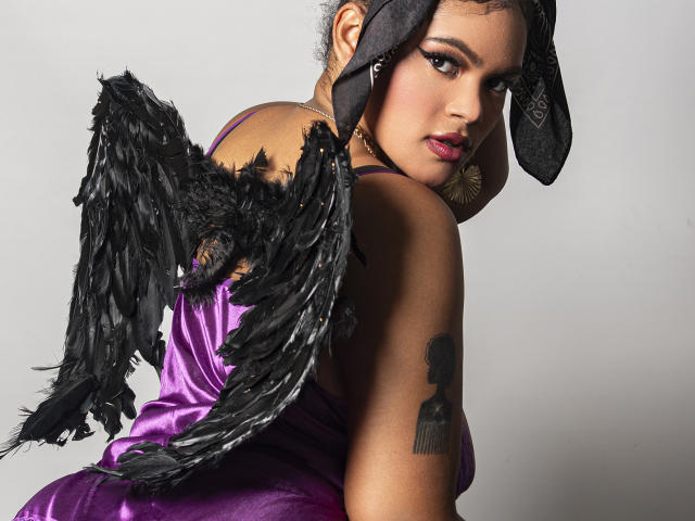 Opera digitale di AliceJelanni con ali nere piumate, abito di seta viola, foulard nero e orecchini a cerchio. Lei sta guardando indietro in modo seducente, rivelando un tatuaggio sulla spalla. Tensione sessuale.