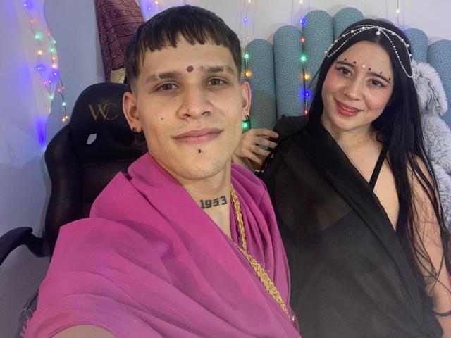 Photographie d'AnjaliRohan, un homme à la chevelure courte, avec un point rouge sur le front, une robe rose, une chaîne en or et un piercing aux lèvres. Assis à côté d'une femme souriante avec de longs cheveux noirs, une robe noire et un point sur le front. Des lumières multicolores en arrière-plan.