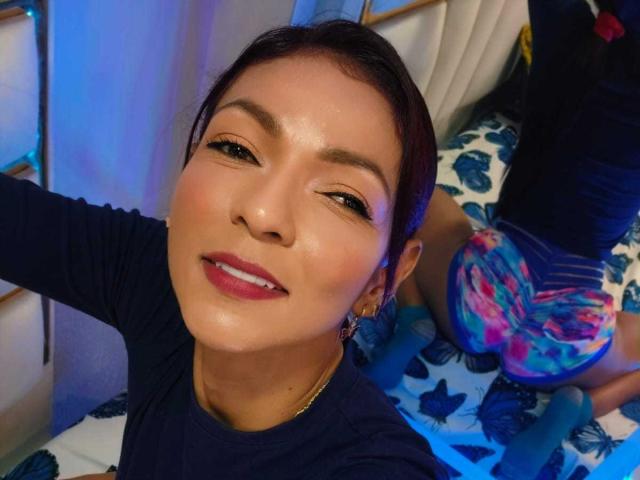 Close-up of HannaPretty, Asian woman with dark hair, wearing a black top, bright red lipstick, and shimmering skin. She's sitting on a bed with blue and black patterned sheets. Vibrant blue light. Fucking sexy.