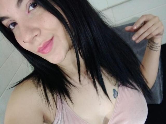 Selfie em close-up de JadeRushh, uma mulher de pele clara com cabelos longos e pretos, camiseta rosa e uma pequena tatuagem abaixo do braço esquerdo. mãmora. Ela tem um sorriso discreto, olhando diretamente para a câmera. A mão dela está apoiada no peito..