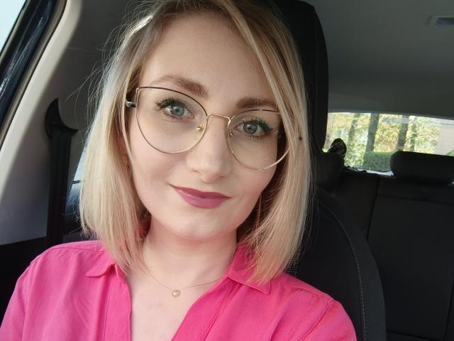 CinderellaMoon, een blonde vrouw met een boblijn, draagt ronde gouden brillen en een felroze shirt, en zit in een auto. Kijkend in de camera met een subtiele glimlach. Heel erg aantrekkelijk.