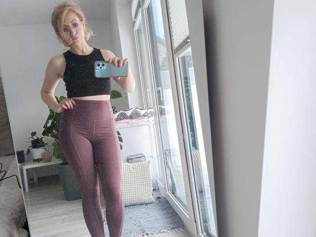 CinderellaMoon se tient dans une pièce lumineuse, en train de prendre un autoportrait avec un téléphone bleu. Elle porte un soutien-gorge de sport noir et des leggings marron ajustés, qui mettent en valeur sa silhouette mince et tonique. La lumière naturelle inonde l'espace..