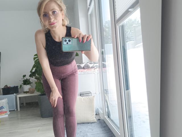 CinderellaMoon macht ein Selfie vor einem Spiegel und lehnt sich dabei nach vorne, wobei sie mit einer Hand auf ihrem Oberschenkel ruht. Sie trägt ein schwarzes Spitzen-Tanktop und marineblaue Leggings, und sie hat eine Brille auf und rote Fingernägel. Helles Sonnenlicht.