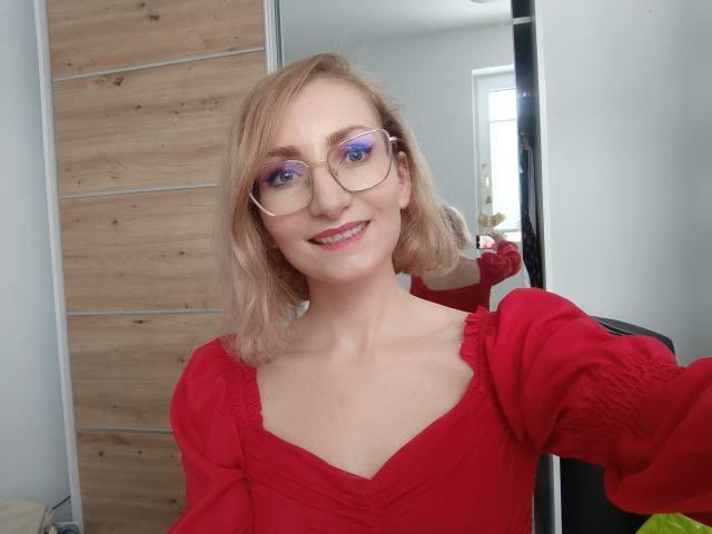 CinderellaMoon, smiling with blue eyes and round glasses, wearing a red dress with puffed sleeves, blonde shoulder-length hair, standing indoors, light wood background, bright light from right. Fucking cute.