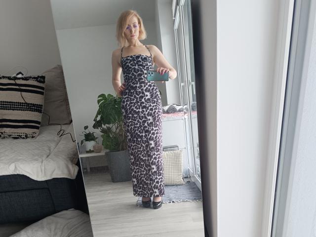 CinderellaMoon staat bij een raam, gekleed in een zwart-wit bloemenjurk, en houdt een groene handdoek vast. Ze heeft blond haar en draagt een bril. Hel licht accentueert haar ronde figuur. Bed en planten op de achtergrond.