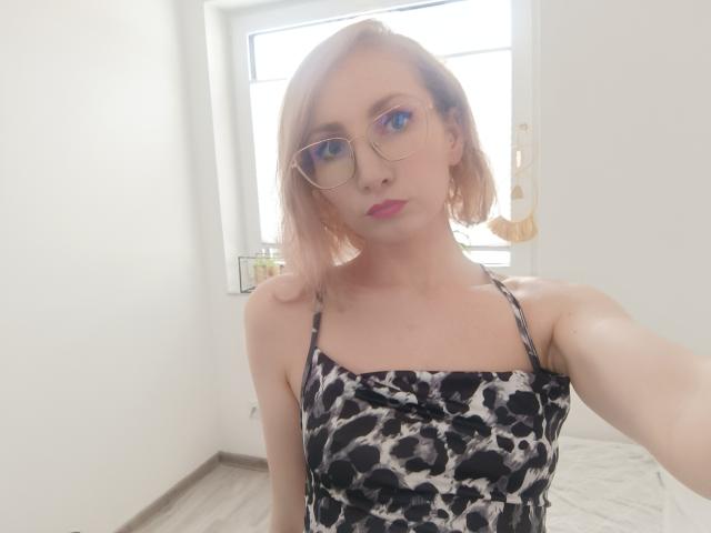 CinderellaMoon, bleekhuidig, blond, met een bril, draagt een jurk met luipaardprint en spaghettibandjes, en staat in een lichte kamer. zonlicht door het raam, zachte uitdrukking, subtiele seksuele spanning, nonchalant selfie, focus op het bovenlichaam, moderne stijl..