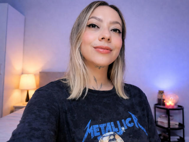 Close-up de KayleeM, uma jovem com cabelo loiro na altura dos ombros, vestindo uma camiseta preta do Metallica, sorrindo levemente. Tatuagens visíveis no pescoço, luz suave no quarto, abajures desfocados ao fundo..