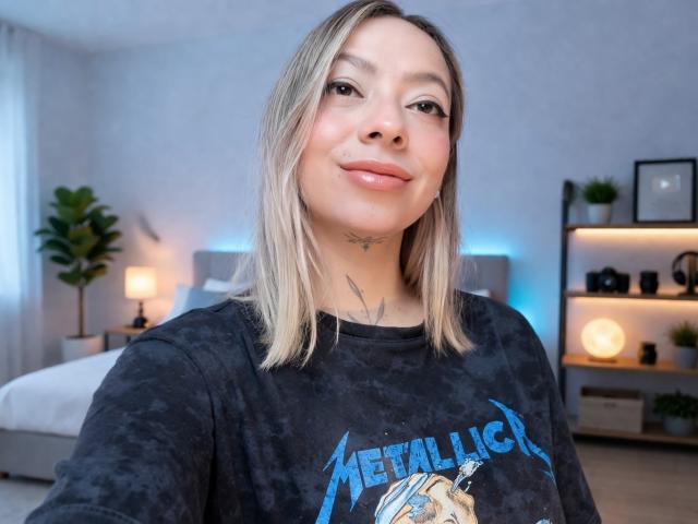 Fotografía de KayleeM, mirando a la cámara, sonriendo ligeramente, vistiendo una camiseta negra de Metallica con letras azules, cabello rubio corto. cabello, pequeña tatuaje en el cuello, fondo de dormitorio con luces azules, plantas y estanterías..