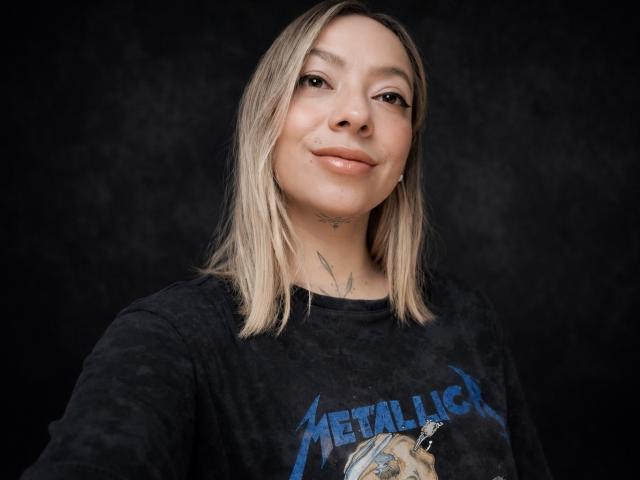 Photograph of KayleeM, light-skinned with blonde shoulder-length hair, wearing a black "Avenged Sevenfold" t-shirt, smiling slightly, visible tattoos on neck, black background, casual, slightly provocative, confident.