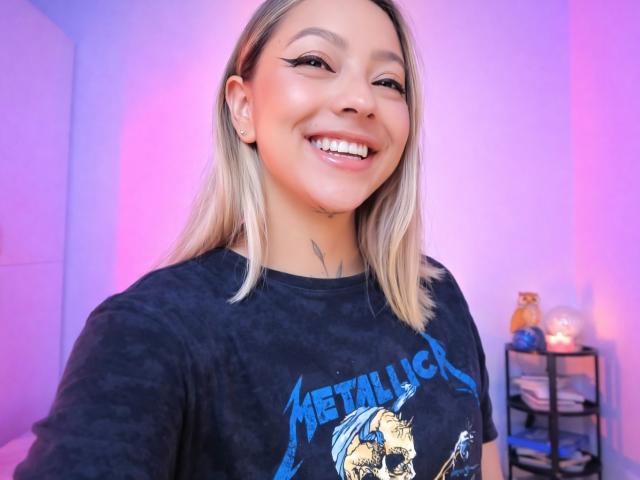 Primer plano de KayleeM sonriendo, mostrando dientes blancos brillantes, y vistiendo una camiseta negra de Metallica. Cabello rubio, piel clara, tatuaje visible en el cuello. Fondo borroso de color morado y rosa. Actitud confiada y con una sonrisa burlona..