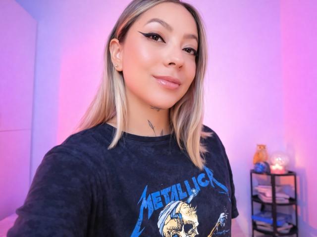 Fotografía de KayleeM, una mujer de piel clara con cabello rubio lacio, vistiendo una camiseta negra de Metallica, con una sonrisa burlona. maquillaje sutil, fondo rosa y morado, estante desordenado con objetos en el fondo..