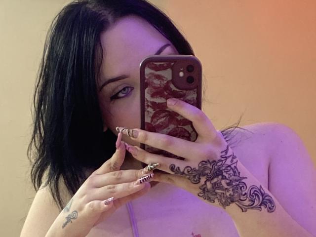 Foto van AlayaWillow, een vrouw met een bleke huid en zwart haar, die een telefoon vasthoudt met een bloemenmotief, waarbij ze haar gezicht bedekt. Ze heeft ingewikkelde zwarte kant tatoeages op haar rechterhand. Verdomd sexy..