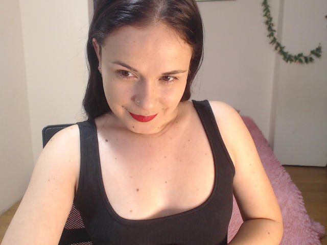 Fotografie von Wendy Schulz mit heller Haut, schwarzen Haaren, rotem Lippenstift und einem schwarzen Tanktop, die mit einem leichten Lächeln sitzt. Dekolleté, weiches, natürliches Licht, rosa, flauschiges Kissen im Hintergrund..