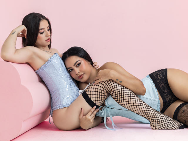 Dos mujeres, PaolaYKatty, están acostadas en un sofá rosa. Una lleva un corpiño de encaje azul claro y medias de red; la otra lleva shorts de encaje negro, medias de red y un corpiño azul. Tensión sexual, abrazo íntimo..