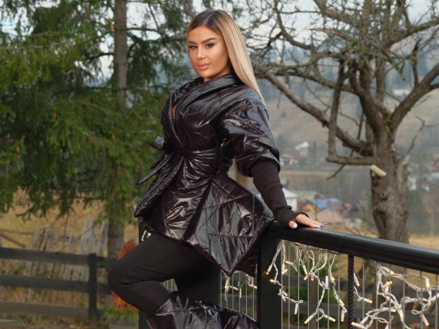 EvaZane leans against a black metal railing, wearing a shiny black dress and black pants. Her long blonde hair falls over her shoulders, with bare trees and string lights in the background. Fucking sexy.