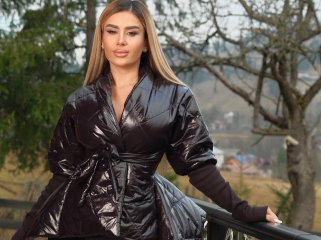 EvaZane se tient fièrement, vêtue d'une veste matelassée noire brillante, ses longs cheveux bruns étant lisses et soignés. Elle est appuyée contre une rambarde noire, avec un paysage boisé en arrière-plan. Extrêmement séduisant..