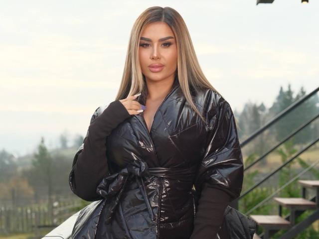 EvaZane, eine attraktive Frau mit langen, glatten, blonden Haaren, die eine glänzende, schwarze, wattierte Jacke trägt, steht draußen und berührt ihre kinn. Hintergrund: Verschwommene Bäume und Treppen. Leicht bewölkter Himmel..