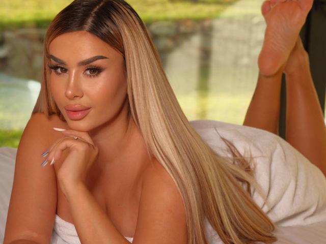 EvaZane, avec ses longs cheveux blonds et lisses, allongée sur un lit, appuyée sur ses coudes, faisant la moue avec ses lèvres pulpeuses, les épaules nues, jambes relevées, pieds croisés, ongles peints en bleu, sexy, séduisant.