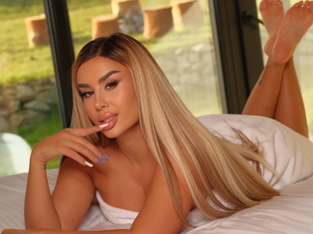 EvaZane ligt op een wit bed, benen omhoog, voeten gekruist, lang blond haar, volle lippen lichtjes open, hand in de buurt van de mond, blauw. nagels, gebruinde huid, sexy, aantrekkelijk, zacht natuurlijk licht, onscherpe achtergrond buiten..