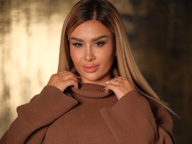 EvaZane, avec ses longs cheveux bruns et lisses, portant un pull col roulé marron, tient le col de ses deux mains. Elle a les lèvres pulpeuses, un maquillage des yeux foncé et un sourire confiant. Une lumière chaude et éclatante..