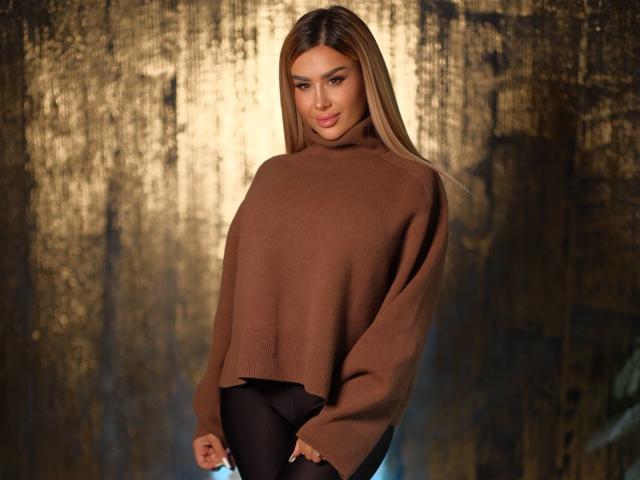 EvaZane, met lang, recht bruin haar, staat voor een donkere, gestructureerde achtergrond met een gloeiend gouden aureool effect. Een bruine, geribbelde, wollen trui met een hoge hals en zwarte broek. Seksuele spanning.