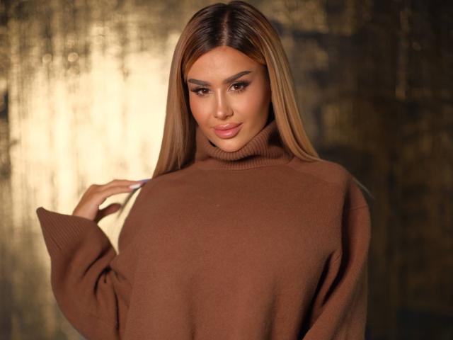 EvaZane, lang bruin haar, bruine coltrui, verleidelijke uitdrukking, hand die de trui aanraakt, warme, lichte achtergrond, zachte focus, sensuele uitstraling, zelfverzekerde pose, lichte glimlach, glanzende lippen, bruine ogen.