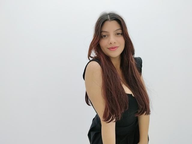 Fotografia di CheryDesire con lunghi capelli castani lisci, che indossa un abito nero e sta in piedi di fronte a uno sfondo bianco. Ha un sorriso delicato, un trucco leggero e un décolleté evidente. Incredibilmente sexy..