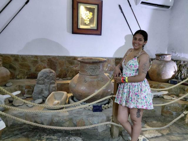 PauletteLoveX stands in a rustic room with large clay pots, wearing a floral dress, smiling, holding a red object. She has tattoos on her arm and thigh, with a sexual tension vibe.