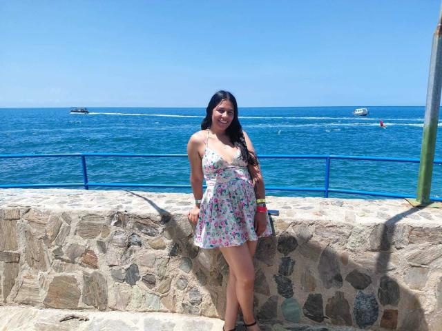 PauletteLoveX está de pie sobre un muro de piedra iluminado por el sol, junto al océano azul, vistiendo un vestido blanco con estampado floral, tacones negros y gafas de sol. una pulsera. Ella se apoya en la pared, con las piernas separadas.