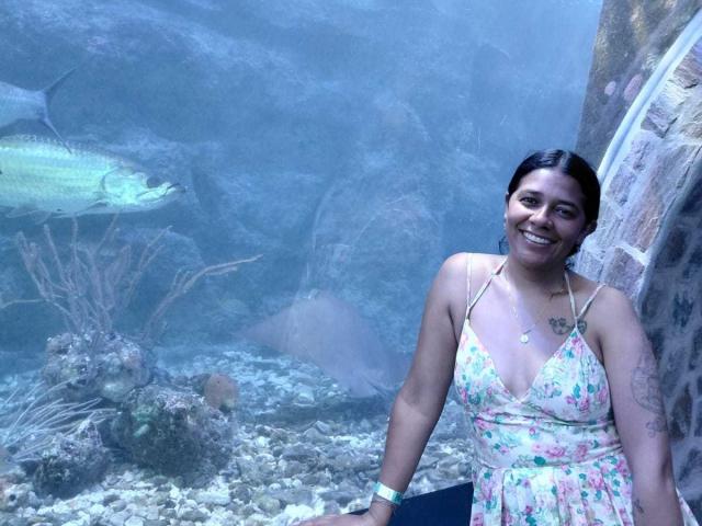 Fotografía de PauletteLoveX con piel de color marrón medio, cabello negro y un vestido floral, de pie frente a un fondo azulado. acuario, sonriendo y con tatuajes visibles en su brazo derecho. Plantas acuáticas en el fondo.