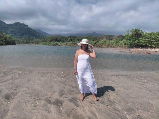 PauletteLoveX está de pie en una playa arenosa, de espaldas a la cámara, vistiendo un vestido blanco sin tirantes y un sombrero de sol. Ella está mirando un lago tranquilo con colinas verdes y un cielo nublado. Muy atractivo..