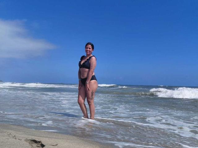 Photograph of PauletteLoveX standing in shallow ocean water, wearing a black bikini, with a clear blue sky and white waves in the background. She has a sexy, confident stance.