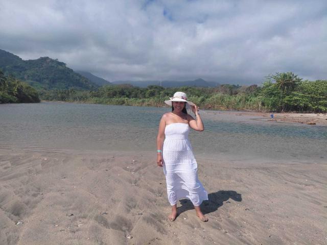 PauletteLoveX, vestida con un vestido blanco sin tirantes y un sombrero blanco, está de pie en la orilla arenosa de un río, sosteniendo su sombrero. Su figura curvilínea y su postura segura evocan tensión sexual. Montañas y cielo nublado en el fondo.