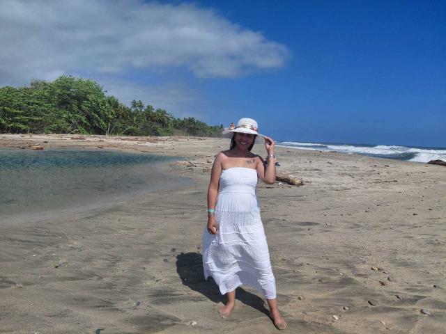 PauletteLoveX, standing on a sandy beach, wearing a white strapless dress and sun hat, barefoot, looking at the camera with a relaxed pose. Clear blue sky, green trees in background.