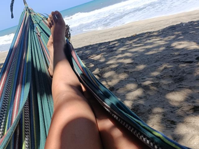 Primer plano de las piernas desnudas de PauletteLoveX, extendidas en una hamaca a rayas de colores en una playa, con los pies colgando y los dedos apuntando. hacia arriba, sombras de árboles cercanos sobre la orilla arenosa, olas del océano en el fondo..