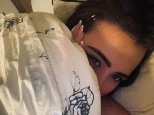 Foto em close-up de AnnyBentleii, com olhos castanhos espiando por baixo de um cobertor branco com estampas florais pretas. Seu cabelo escuro está ligeiramente despenteado, e ela está olhando diretamente para a câmera. A mão dela está próxima do rosto..