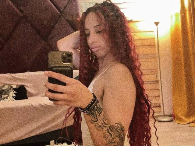 Nymera with long red curly hair, white tank top, tattooed arm, holding phone, taking mirror selfie, looking down, in dimly lit room, yellow curtain in background.