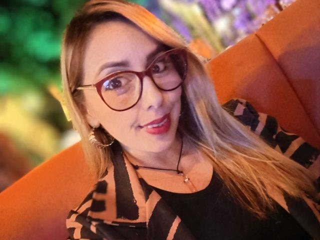 Close-up of CaroLarson with long blonde hair, wearing brown-tinted glasses and a black and orange patterned top. She has a warm smile, red lipstick, and gold hoop earrings. The orange cushion she's sitting on contrasts with her light skin. The blurred background has green and purple hues. The image has a slightly warm, soft-focus effect. CaroLarson's makeup is subtle, highlighting her natural features. Her hair is straight and falls to the side.