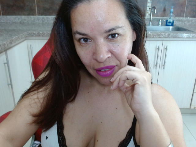 Close-up photo of LizethBells. She has medium skin, brown hair, and bright purple lipstick. She's wearing a black lace bralette, showing cleavage. Her right hand touches her chin. She's sitting in a red chair in a kitchen with white cabinets and a granite countertop. The background includes a sink with a blue bottle. Her expression is slightly amused, and she has small freckles on her face. The image has a casual, selfie-style composition.
