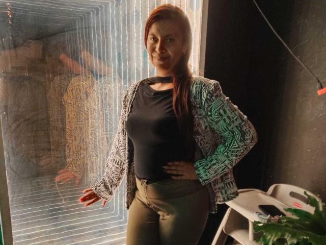 AmbeRouse with red hair, black top, and green pants, wearing a patterned cardigan, standing with hand on hip, reflection in a patterned glass panel, smiling.