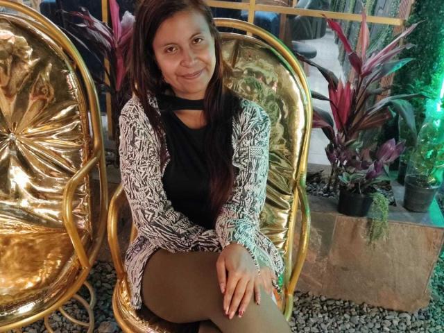 AmbeRouse with brown hair, black top, and white lace cardigan, sitting on a gold chair, smiling, showing her brown tits and ass, with green plants in the background.