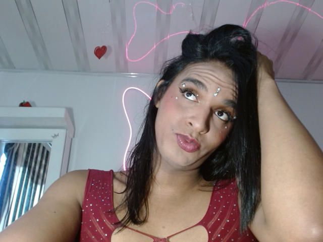  IdiiaLatina has medium brown skin and dark, straight hair, pulling it up with her right hand. She wears a red, sheer top with a keyhole cutout, showing a bit of cleavage. Her makeup includes small, white dots on her cheeks and a silver nose stud. She has a pink nose and slightly pouty lips. A pink neon light and a small red heart are on the white, wooden ceiling. A window with vertical blinds is on the left. The image has a casual, selfie-style composition.