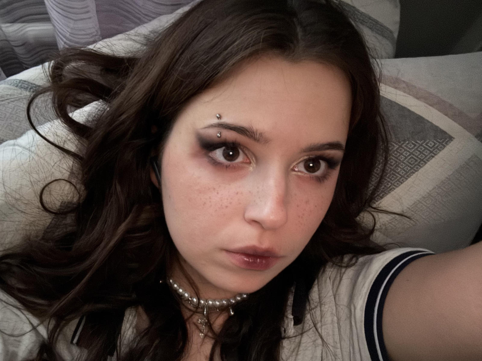 KelsieWoolcock has a face piercing above her right eyebrow, dark wavy hair, brown eyes, freckles, and wears a pearl choker with a necklace. She's looking at the camera.