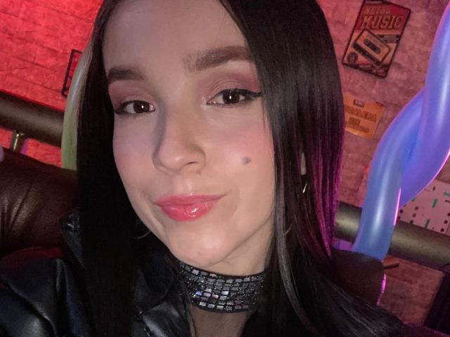 MeliLopezX with black hair, pink eyeshadow, and glossy pink lips, wearing a black choker and leather jacket, has a small mole on her left cheek.
