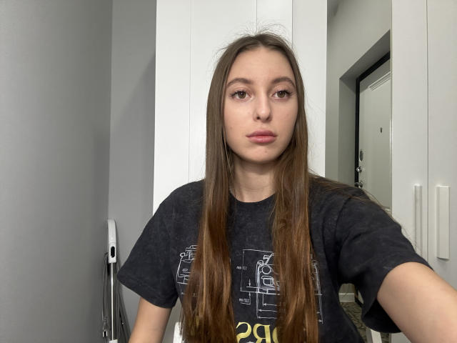 ErinMarrujo with long brown hair, wearing a black t-shirt, has a neutral expression, brown eyes, and is holding a phone for a selfie. Her tits are small.
