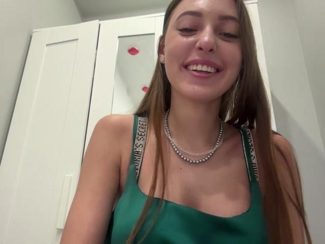 ErinMarrujo with long brown hair, green tank top, and silver necklace, smiling at camera, showing her tits, wearing "VANESSA SANTO" straps on tank top, in room.