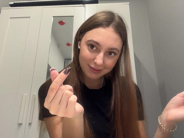 ErinMarrujo, avec de longs cheveux bruns, un t-shirt noir et une peau pâle, arborant des ongles peints en violet, souriant légèrement, portant des écouteurs blancs et un bracelet, dans une pièce grise..